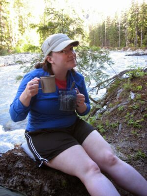 A woman with two mugs sitting on a rock next to a river