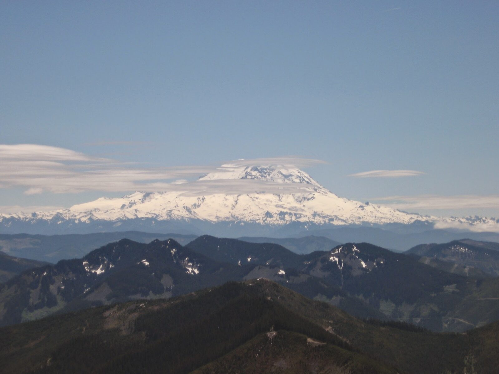 Mt Rainier, a high snow covered mountain, in the background surrounded by a few clouds on an otherwise sunny day. Smaller mountains are in the foreground from Bandera Mountain, one of the best hikes in Washington