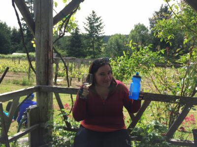 A woman is smiling on a patio in a vineyard. She is holding a wine glass and a biking waterbottle
