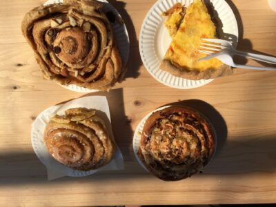 There are four pastries on a picnic table outside in the sun. There are three cinnamon rolls and one quiche with a fork