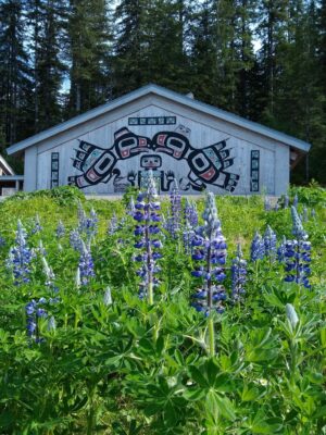 A wooden building with Tlingit art on the side. Purple wildflowers are in the foreground