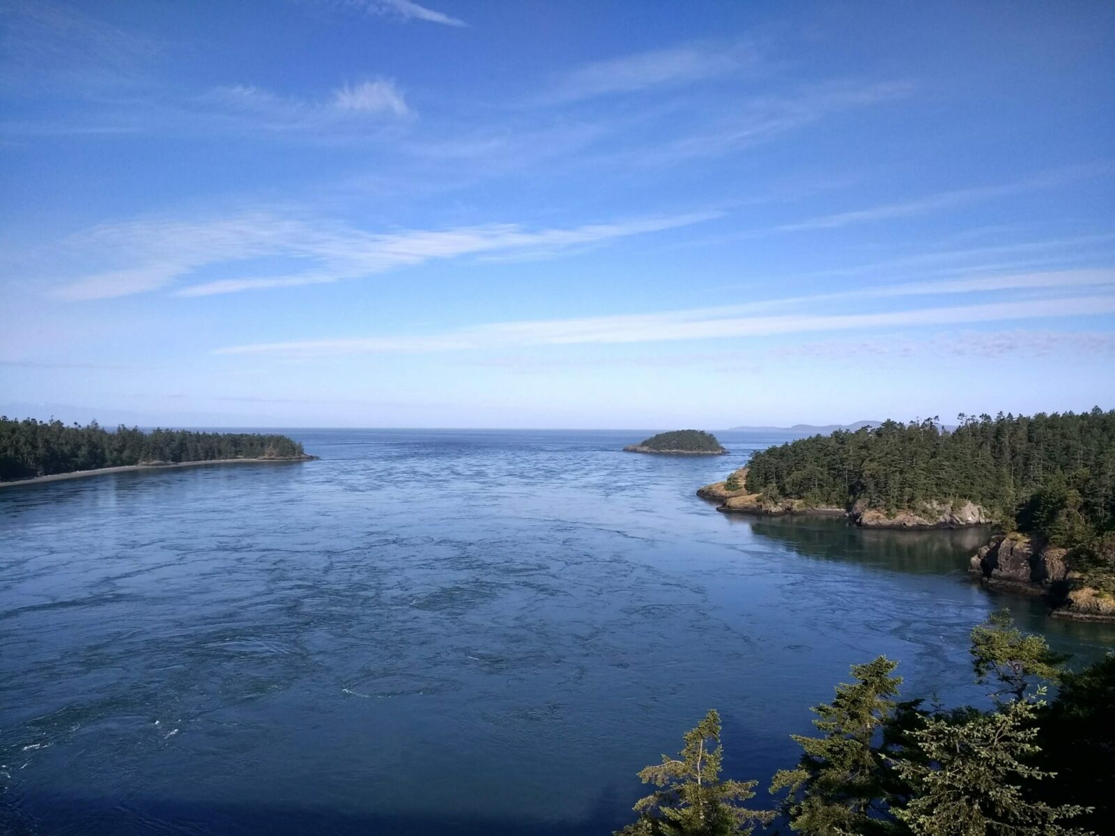 A view from a high bridge on a sunny day. There is churning water below and forested hillsides on both sides as well as islands in the distance