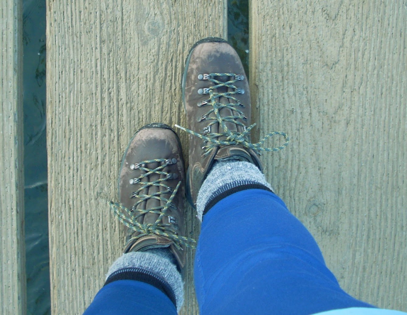 Two worn hiking boots seen from the person wearing them. A person's lower legs with blue leggings and gray and blue socks are also visible. The person is standing on a wooden path