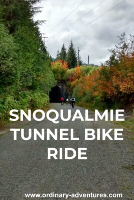 Three cyclists in the distance approaching the entrance to a tunnel on a gravel trail surrounded by green and orange trees with fall color