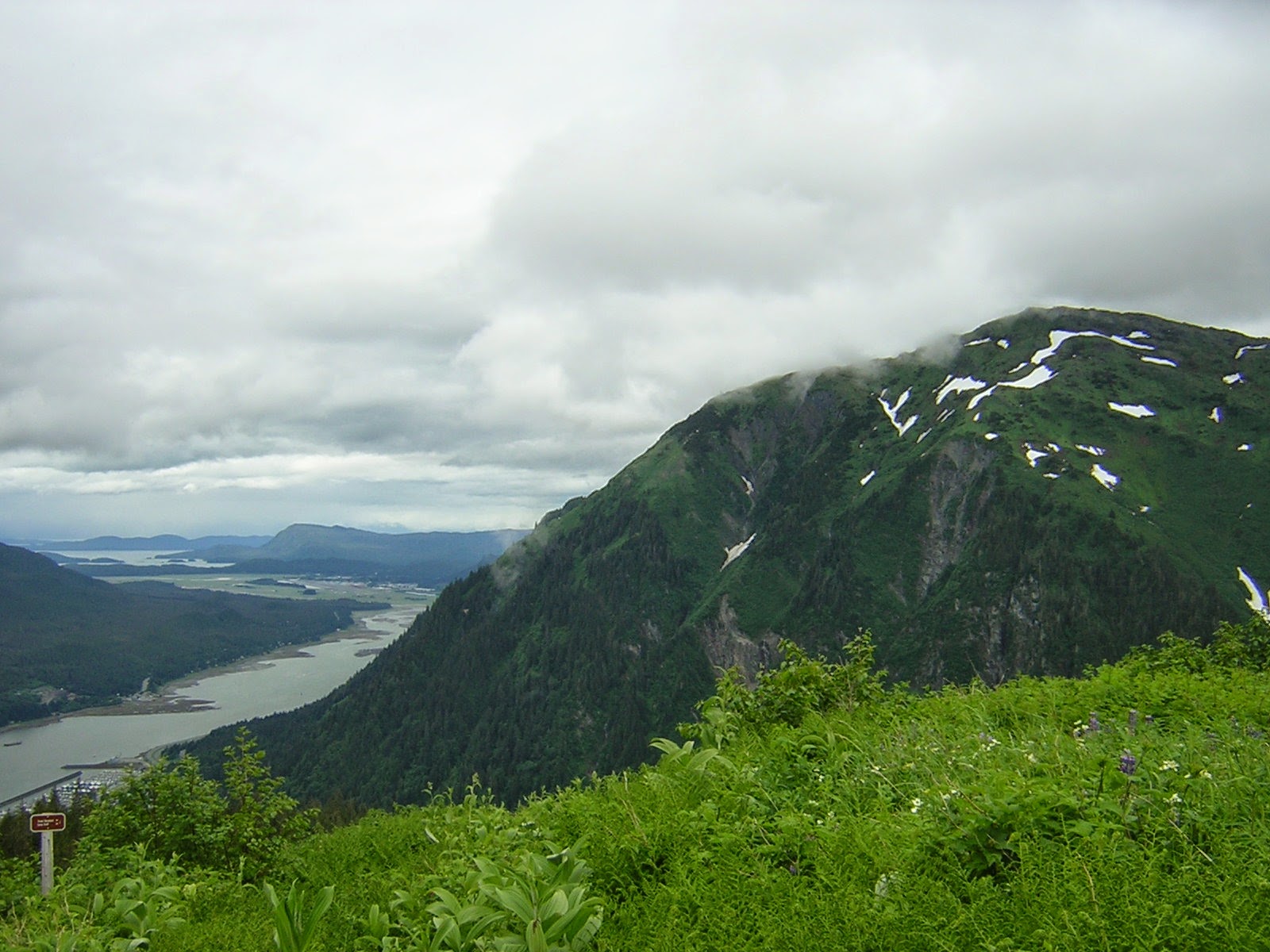 A channel of water between two mountains. There are more mountains in the distance. In the foreground is a green meadow looking down on the water and town below