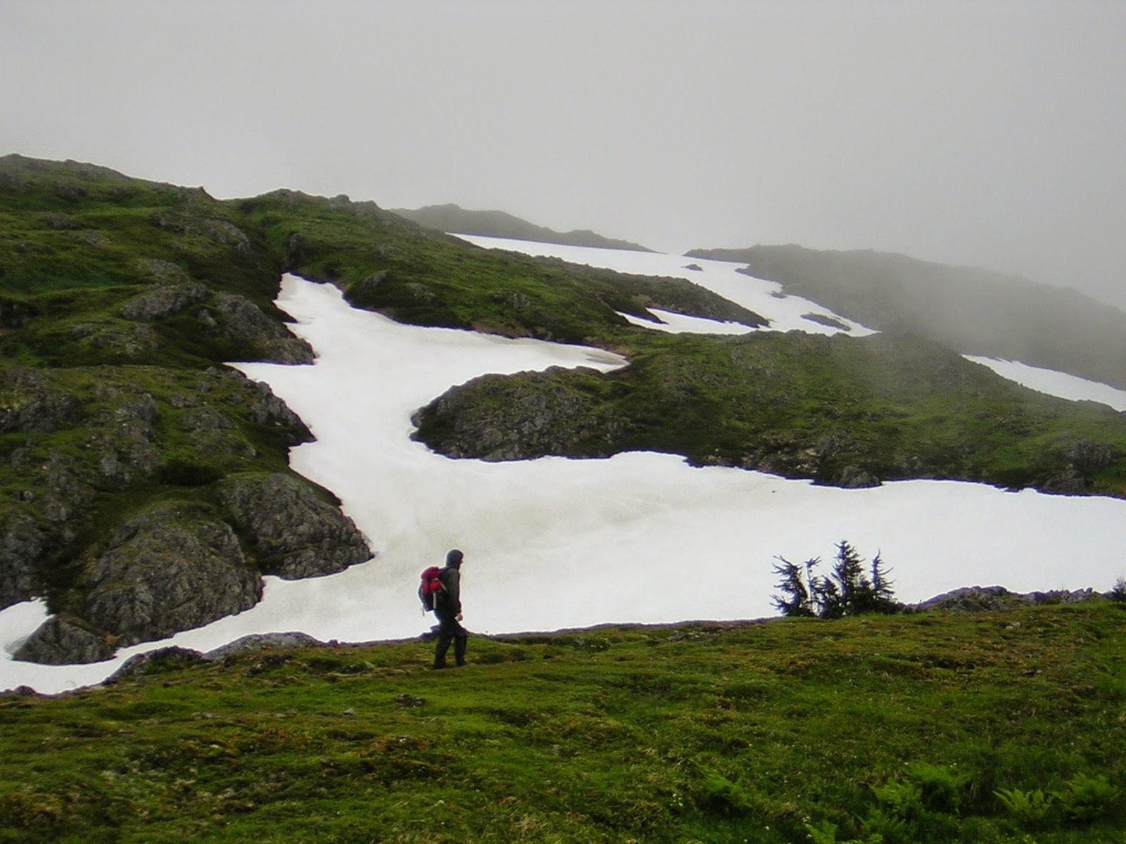 Patches of snow cover a rocky mountain on a foggy day. A person is standing on a grassy area walking across it