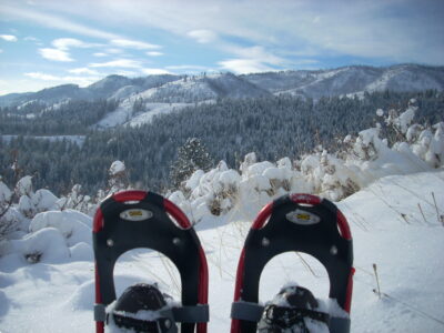 Two snowshoes in the foreground with a snowy landscape in the background. There are snow covered forested hills in the background and a blue sky