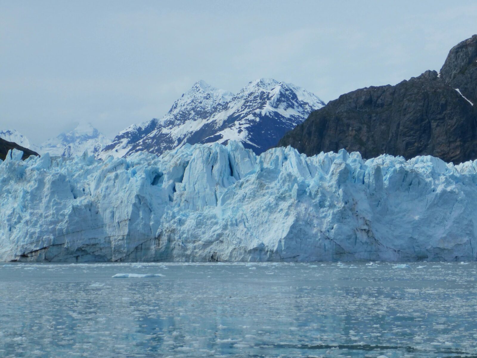 A blue tidewater glacier at the edge of the water. There is lots of ice flating in the water. In the background there are snow covered mountains on a cloudy day
