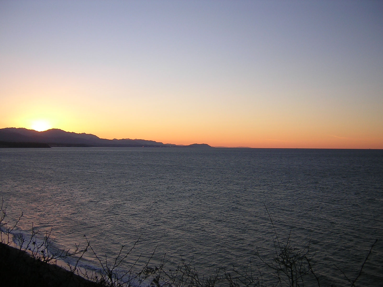 A winter sunset at the Dungeness Wildlife Refuge. The sun has just set behind the mountains and the water is dark gray. There are bushes in the foreground