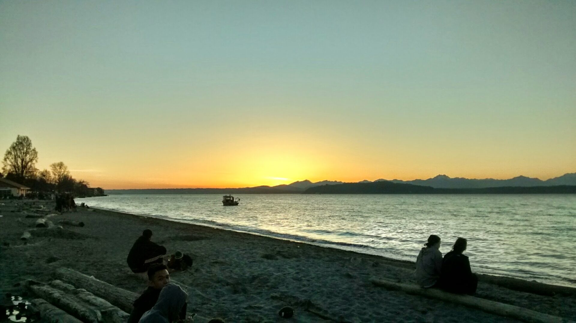 Sunset behind the mountains at a Washington beach, Alki Beach in Seattle. It is getting dark and there are people sitting on logs on the beach, watching the sunset. There is a boat in the water