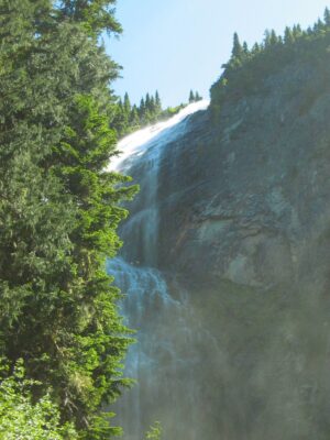 A wide, misty waterfall comes over a rock face with some evergreen trees around it