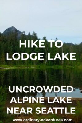 An alpine lake surrounded by evergreen trees. There are a couple of higher mountains visible in the distance. Text reads: hike to Lodge Lake uncrowded alpine lake near Seattle