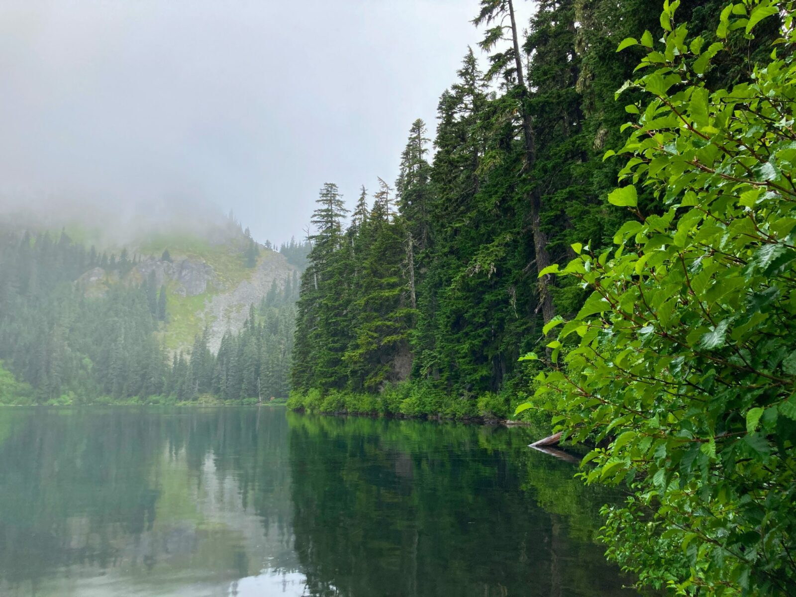 Mirror Lake on a cloudy day is ringed by evergreen trees and mountains which area hidden behind the clouds