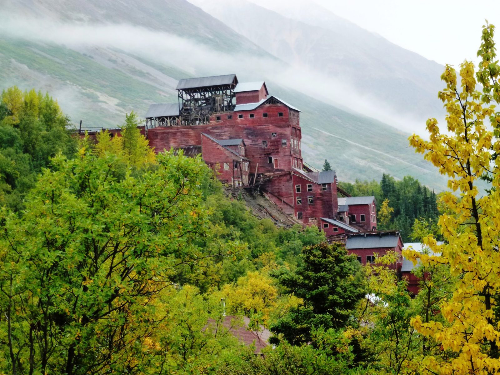 A ruined large wooden structure, the historic Kennicott mine is built into a hillside. The building is painted red with a black roof. There are trees in the foreground and a mountain in the background in Wrangell-St Elias National Park in Alaska
