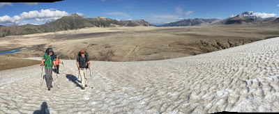 Three hikers walk up a snow field with a volcanic landscape in the background covered in pumice. There are mountains in the distance in the valley of 10,000 smokes in Katmai national park in alaska