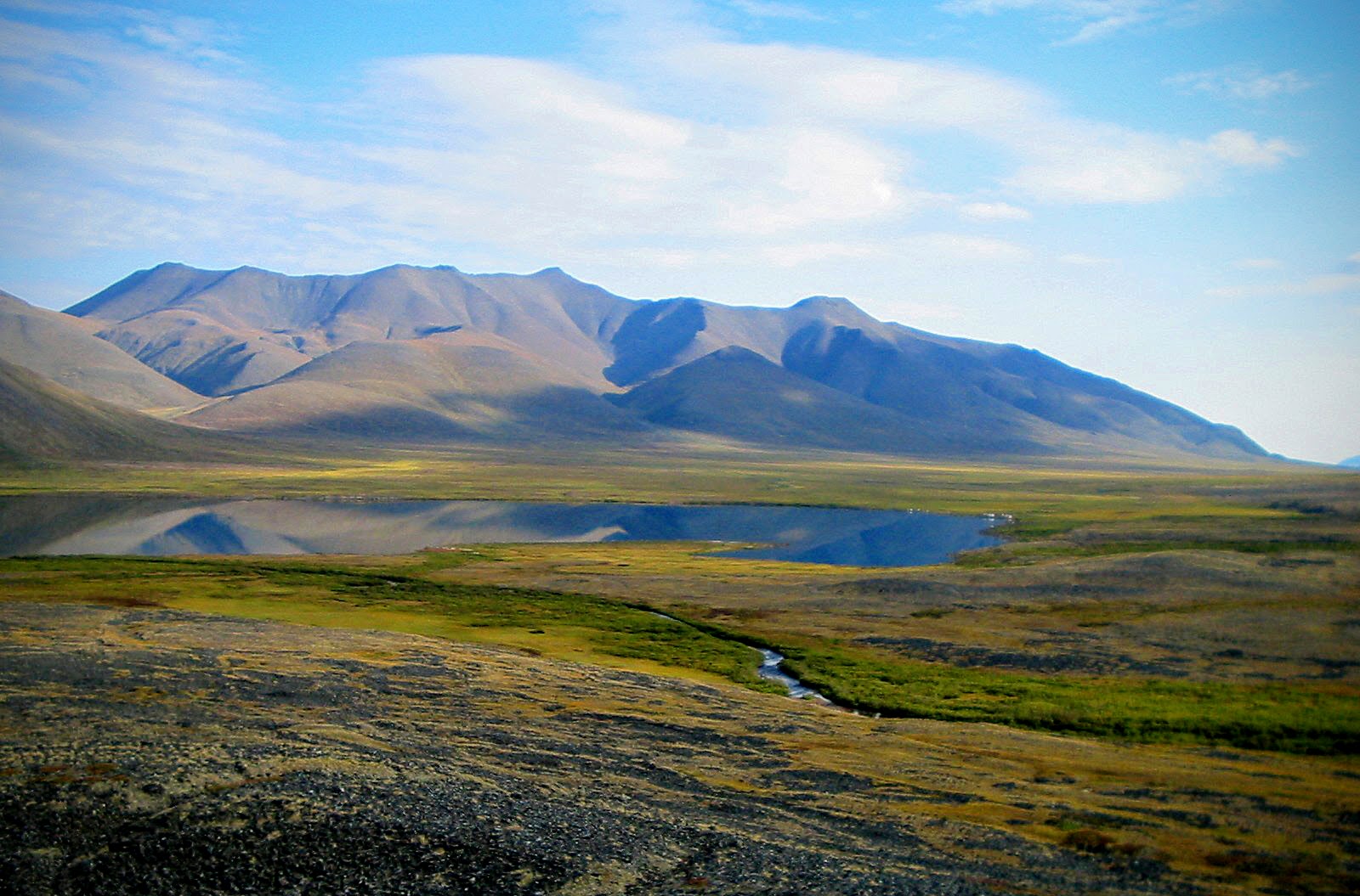 A small, smooth lake reflects brown mountains and green bushes far above the arctic circle in Gates of the Arctic National Park in Alaska.