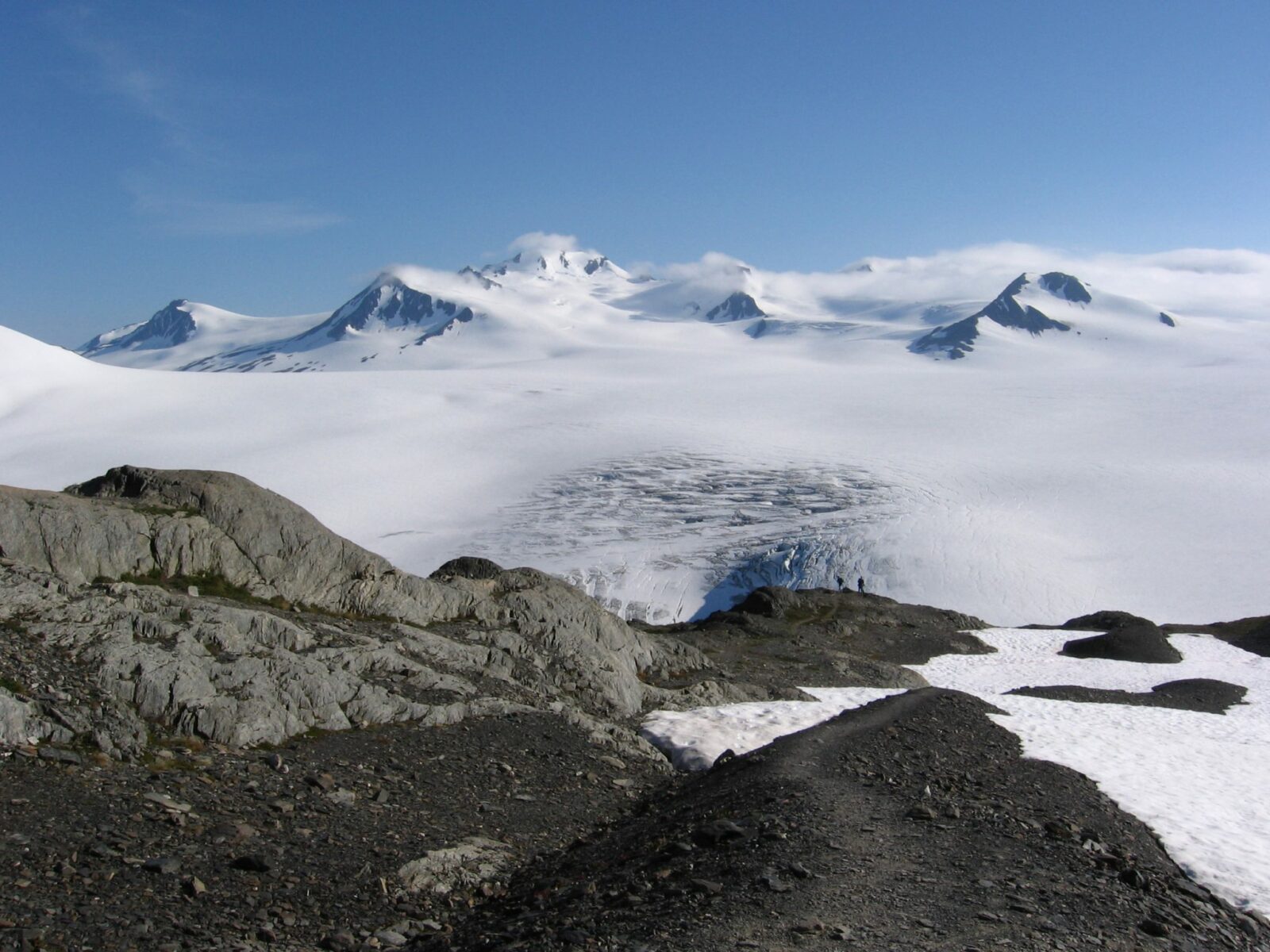 A highlight of Alaska national parks is the Harding Icefield hike. Two hikers in the distance near rocks and a snow field. A glacier in the distance and mountains partially covered in clouds.