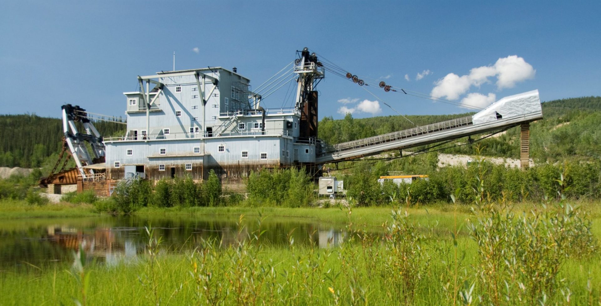 A large gold dredge in a green field with a pond and mountains in the background. The dredge looks like a 5 story metal building with a long arm sticking out one side and more machinery in the back