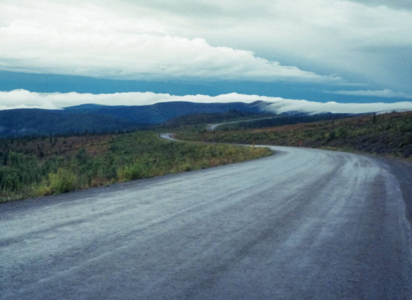 A gravel road winds along an open ridge surrounded by shrubs and distant mountains. It's a rainy day