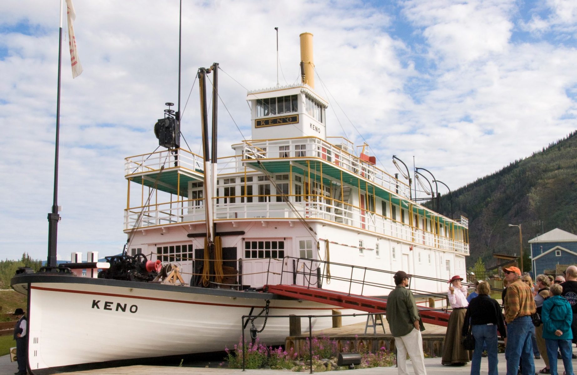 A restored paddle wheeler in downtown Dawson City. It's white with red and green trim and called the Keno. A group of people on a tour are standing in front of it