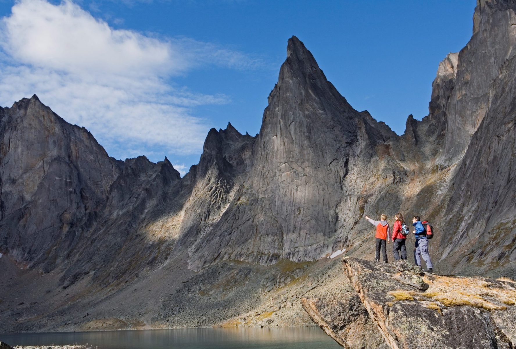 Three hikers stand on a rock next to a lake surrounded by high jagged mountains in Tombstone Territorial Park