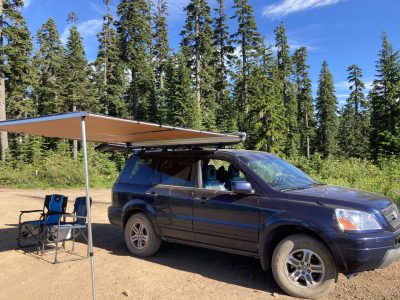 A blue SUV parked in the forest with an awning extended from the roof. There are two camp chairs parked under it