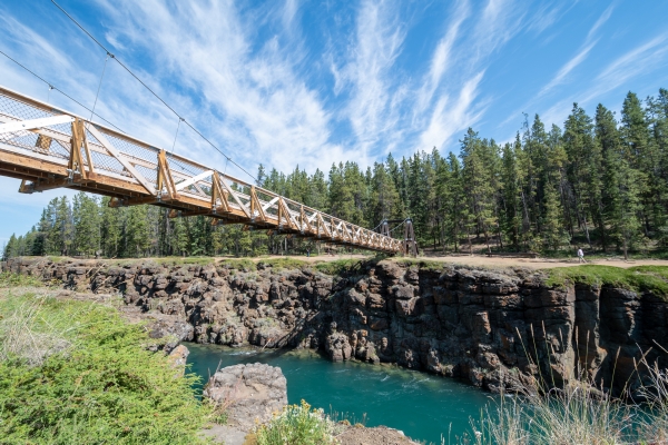 A canyon with steep rocky sides and blue water with a wooden and steel suspension bridge over it. There are forests surrounding the canyon on a sunny day